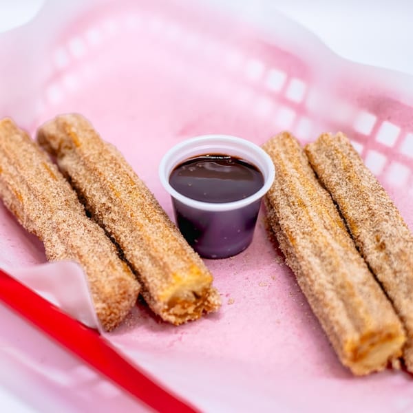 Four cinnamon sugar churros and a small cup of chocolate dipping sauce in a pink basket.