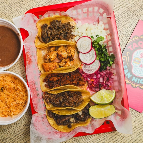 A red basket holds five Taqueria San Miguel tacos with sides of rice, beans, and limes.