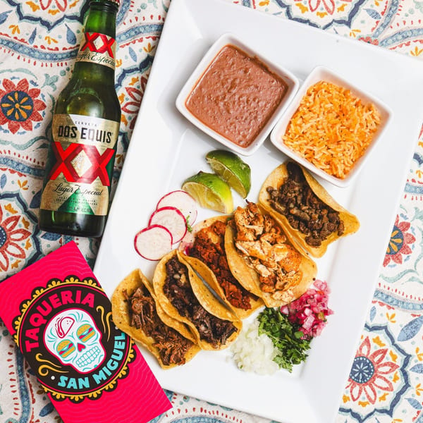 A plate of Taqueria San Miguel tacos with rice, beans, lime, radishes, and a Dos Equis beer.